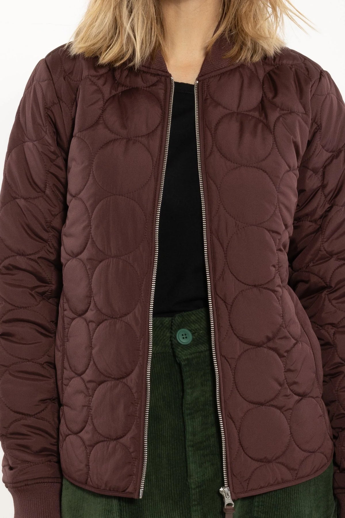 A close-up of a woman wearing a dark red quilted jacket from Danefae with a circular bubble pattern, styled over a black top and green corduroy trousers.