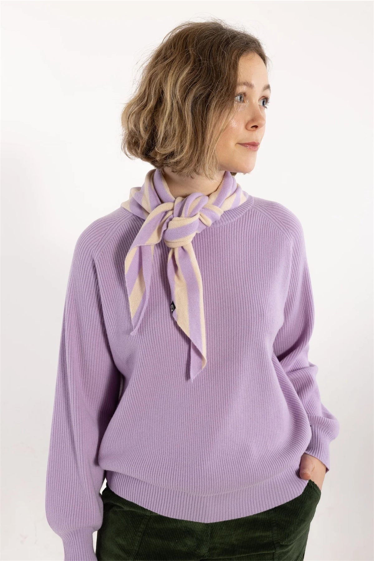 A woman wearing a lavender ribbed knit sweater with a matching lavender and cream striped neck scarf by Danefae tied in a knot, paired with dark green corduroy pants, against a plain white studio background.