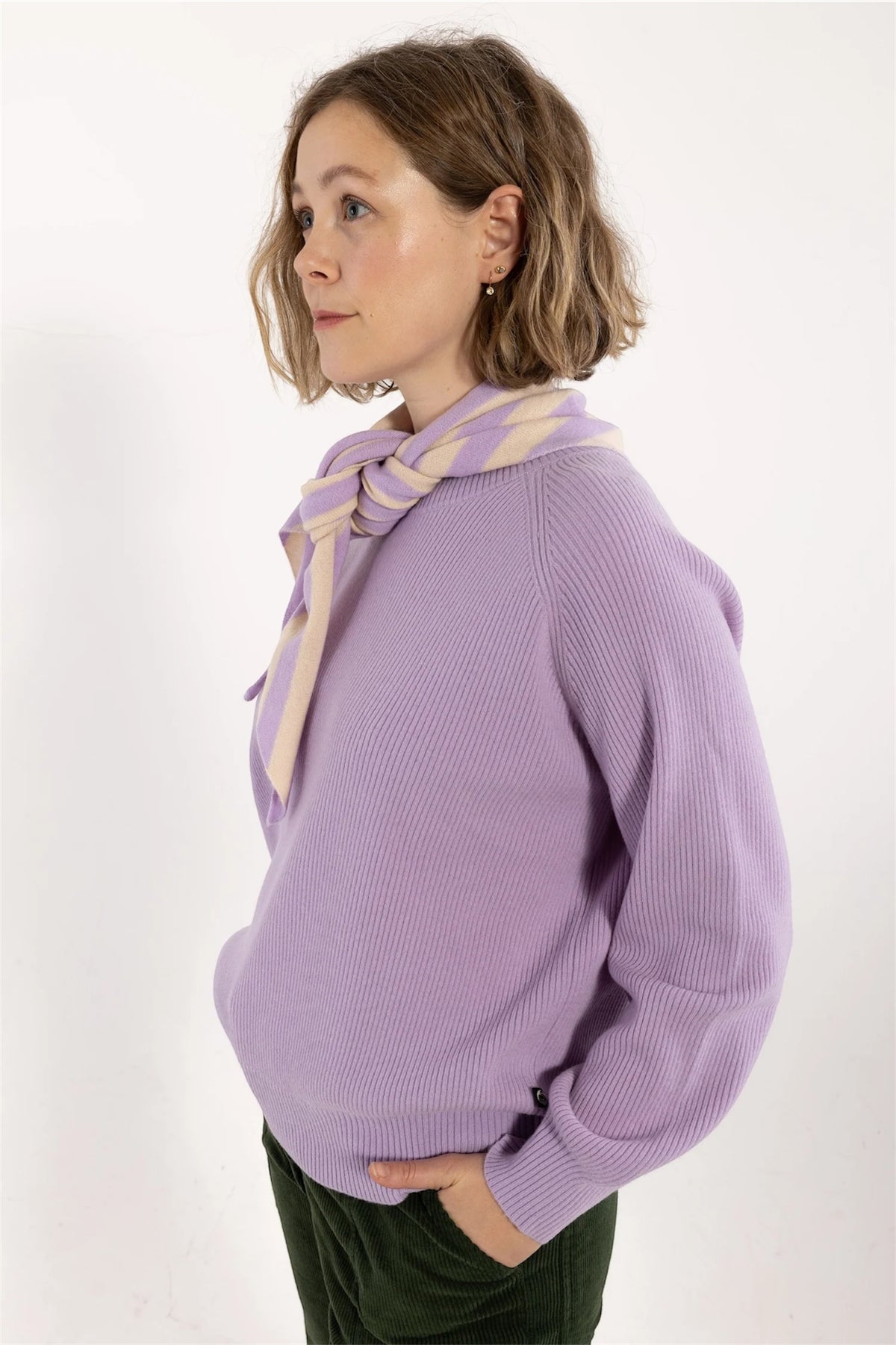 A woman wearing a lavender ribbed knit sweater with a matching lavender and beige striped neck scarf by Danefae, paired with dark green trousers, photographed against a plain white studio background.