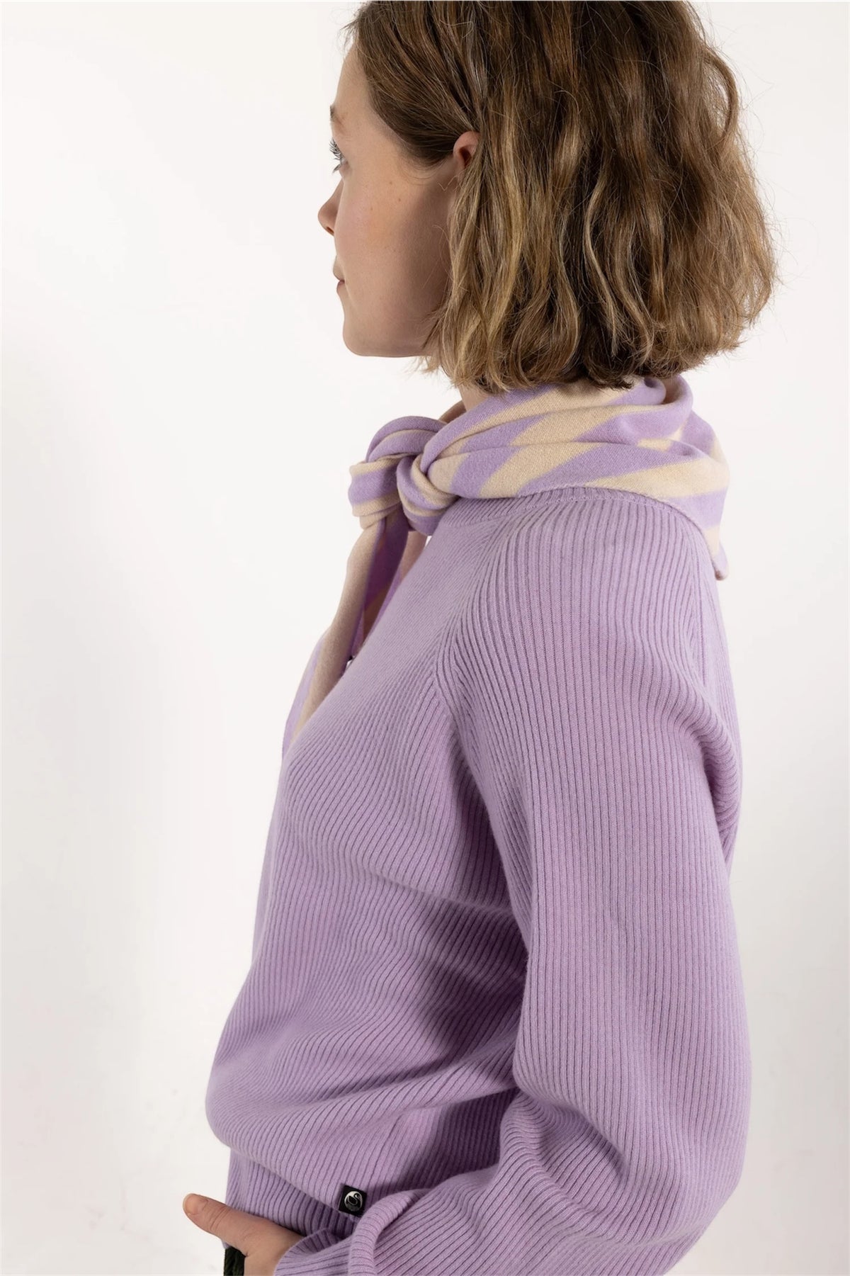 A woman shown in profile wearing a lavender ribbed knit sweater and a matching lavender and cream scarf by Danefae, photographed against a plain white studio background.