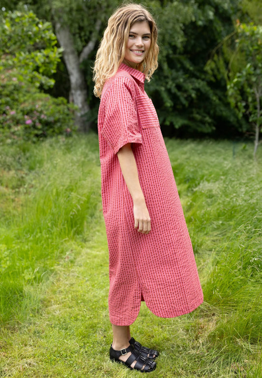 Woman wearing a pink dress standing in a grassy outdoor area with greenery.