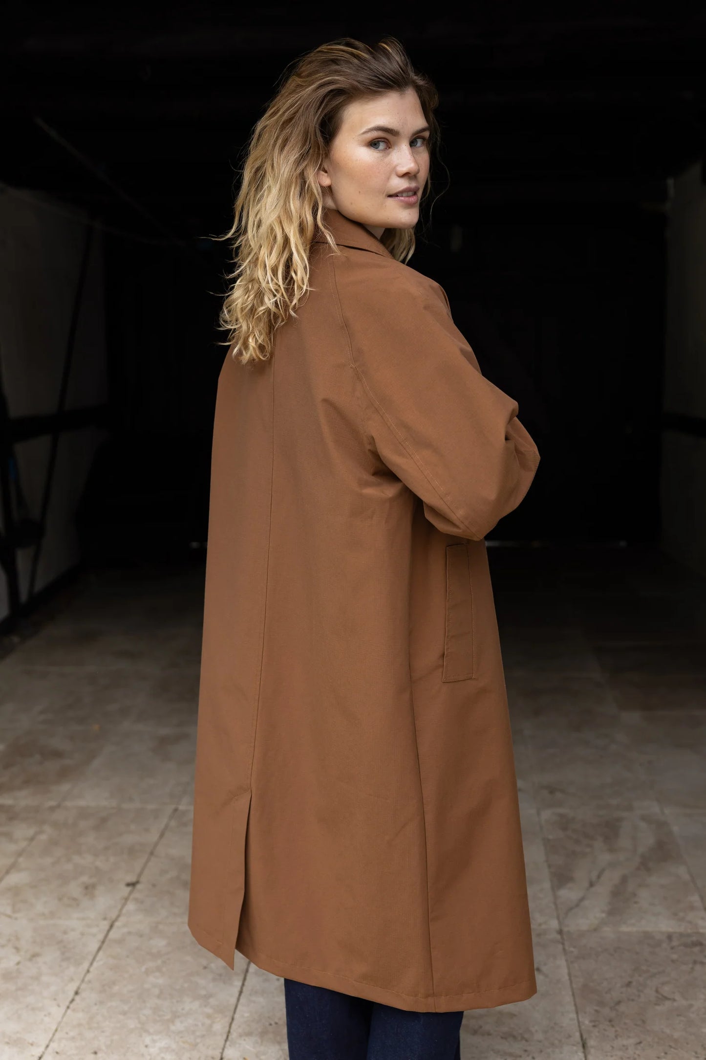 Woman wearing a brown coat standing in a dimly lit room.