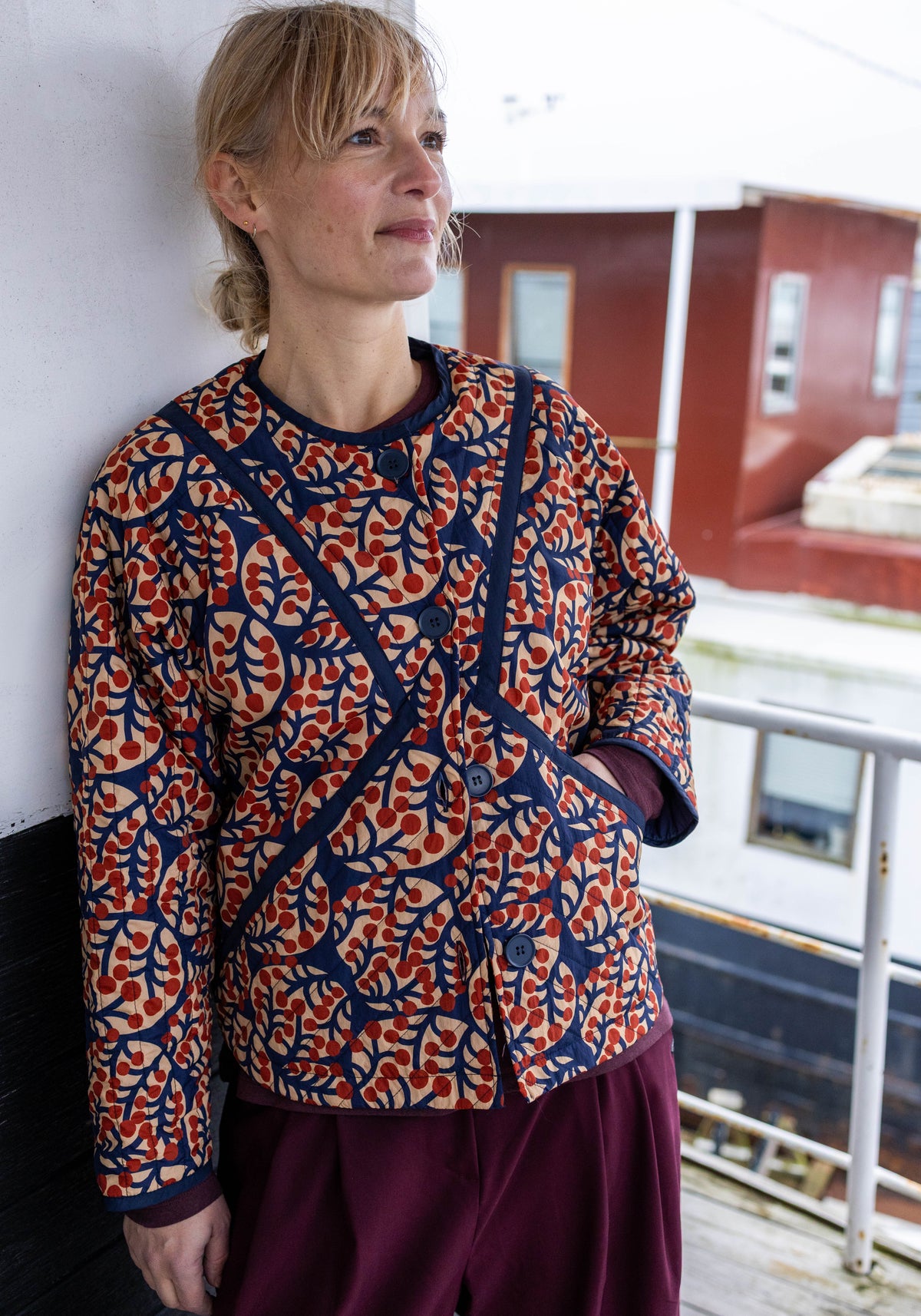 Woman wearing a patterned quilted jacket by Danefae and purple pants standing indoors.