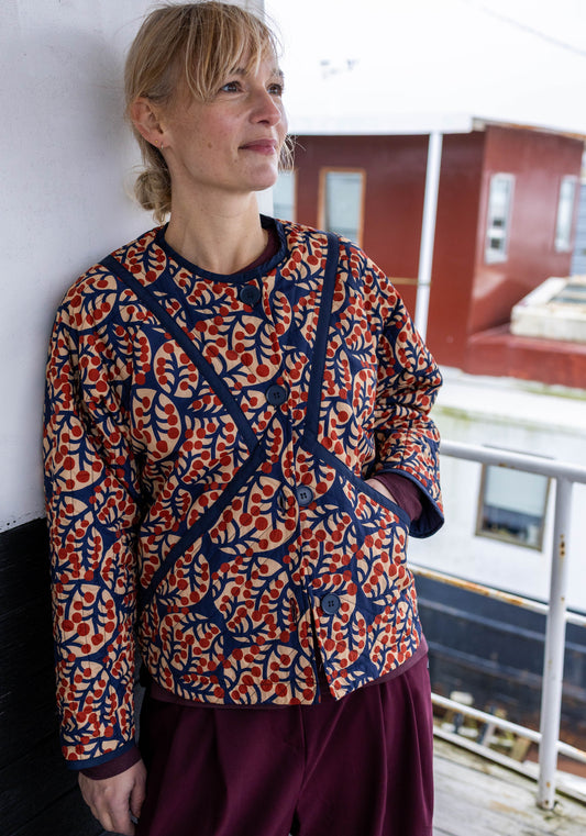 Woman wearing a patterned quilted jacket by Danefae and purple pants standing indoors.