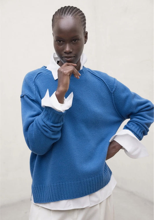 A woman wearing a woolen royal blue crewneck jumper by Ecoalfr layered over a white collared shirt, set against a plain light beige wall. 