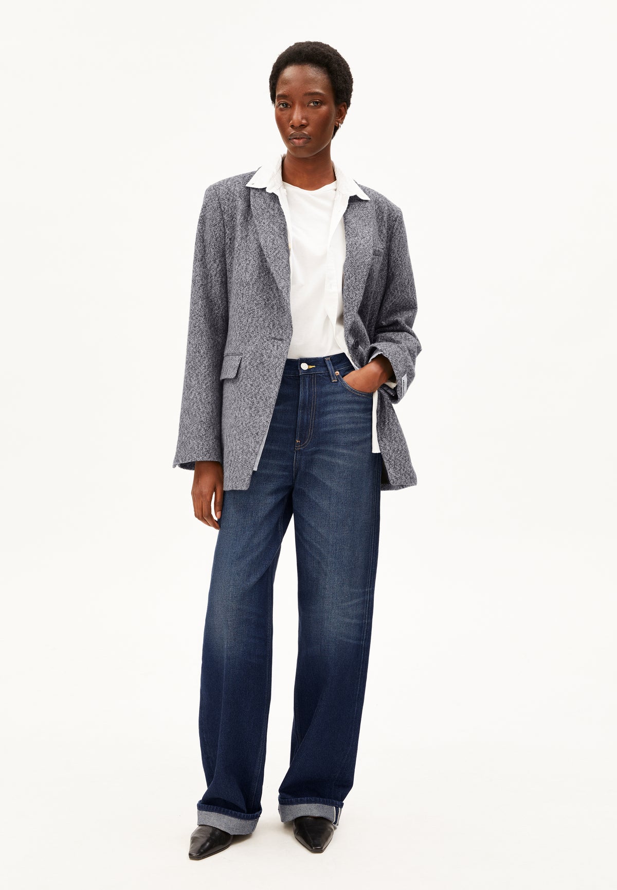 A model stands against a plain white studio background wearing a gray textured blazer over a white collared shirt, paired with dark blue wide-leg Enijaa jeans by Armedangels and black pointed shoes.