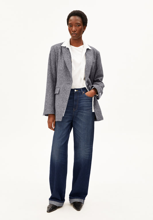 A model stands against a plain white studio background wearing a gray textured blazer over a white collared shirt, paired with dark blue wide-leg Enijaa jeans by Armedangels and black pointed shoes.
