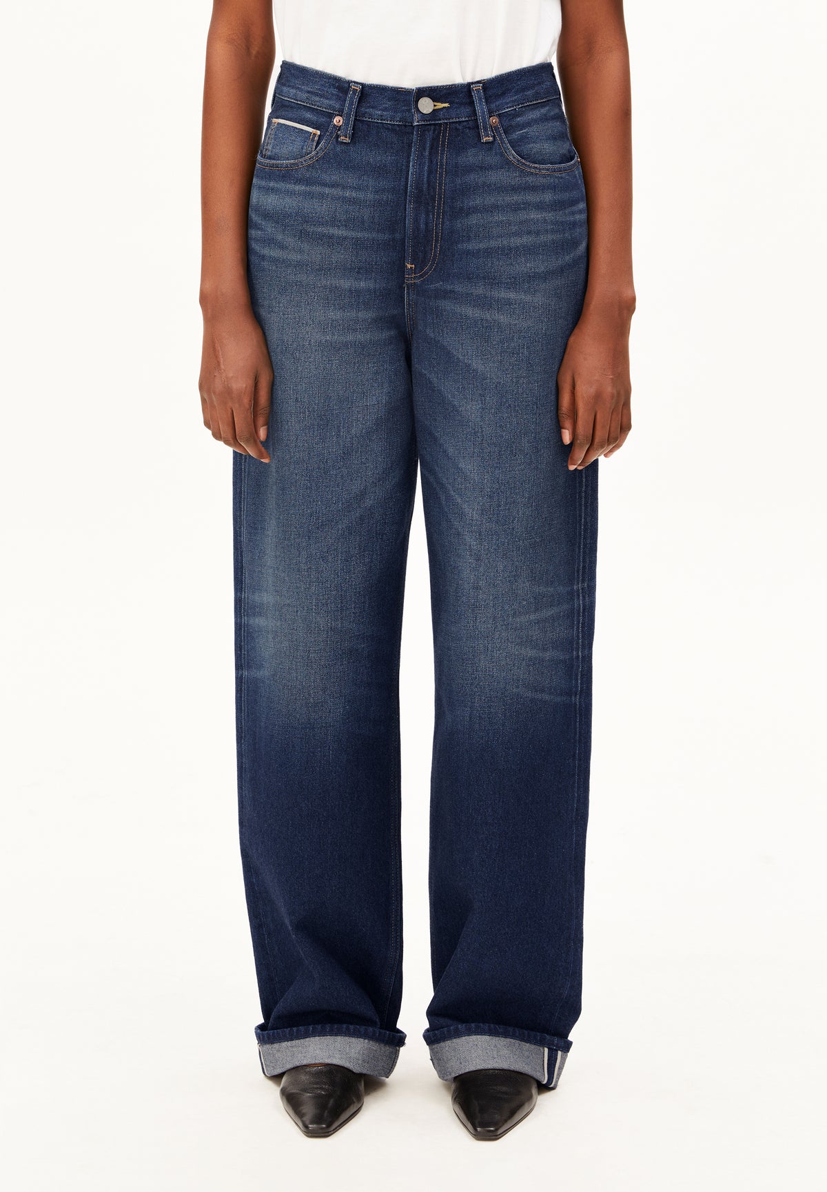 A model wearing a pair of dark blue wide-leg Enijaa jeans by Armedangels with a faded wash, styled with a white top and black pointed-toe heels against a plain white background.