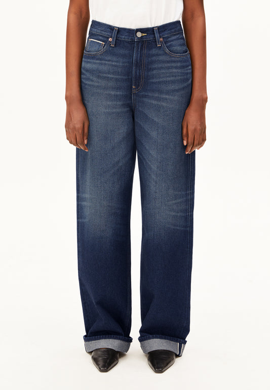 A model wearing a pair of dark blue wide-leg Enijaa jeans by Armedangels with a faded wash, styled with a white top and black pointed-toe heels against a plain white background.
