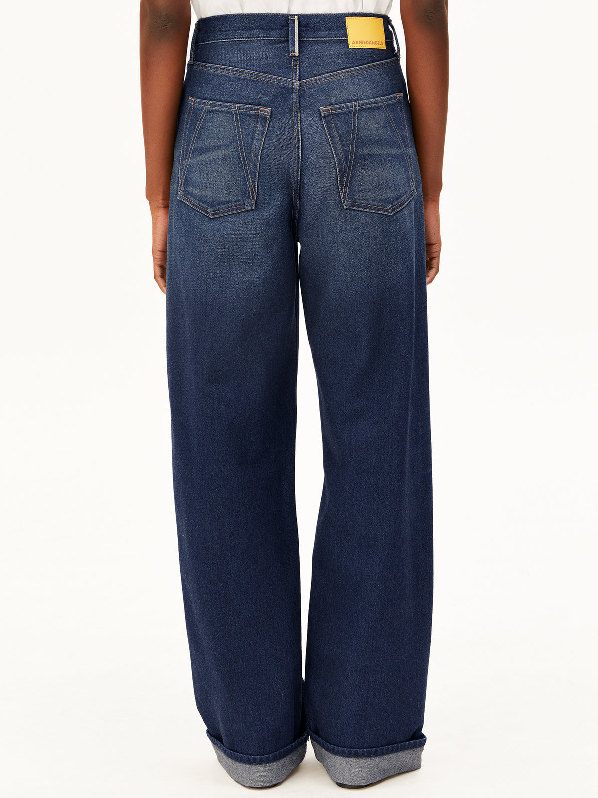 Rear view of a person wearing dark blue wide-leg Enijaa jeans by Armedangels with a yellow brand patch at the back waistband, shown against a plain white studio background.