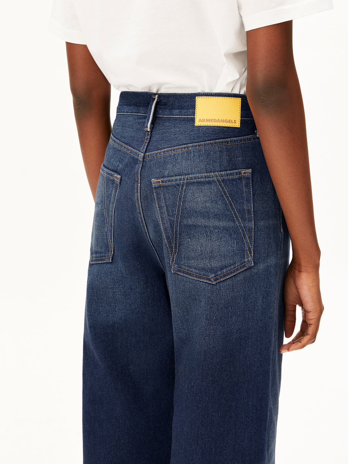 Rear view of a model wearing dark blue wide-leg Enijaa jeans by Armedangels with a yellow brand patch at the back waistband, paired with a white t-shirt against a white studio background.