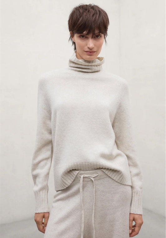 A woman wearing a cream-colored woolen turtleneck jumper from Ecoalf and matching light beige knit pants, standing against a neutral light gray wall.