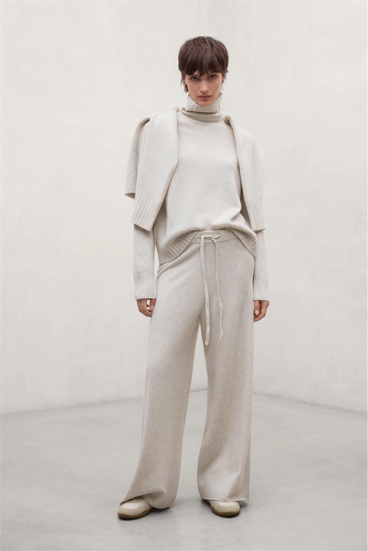 A woman wearing a light beige knit three-piece outfit consisting of a turtleneck sweater, draped cape-style cardigan, and wide-leg drawstring trousers, standing against a neutral light gray wall.