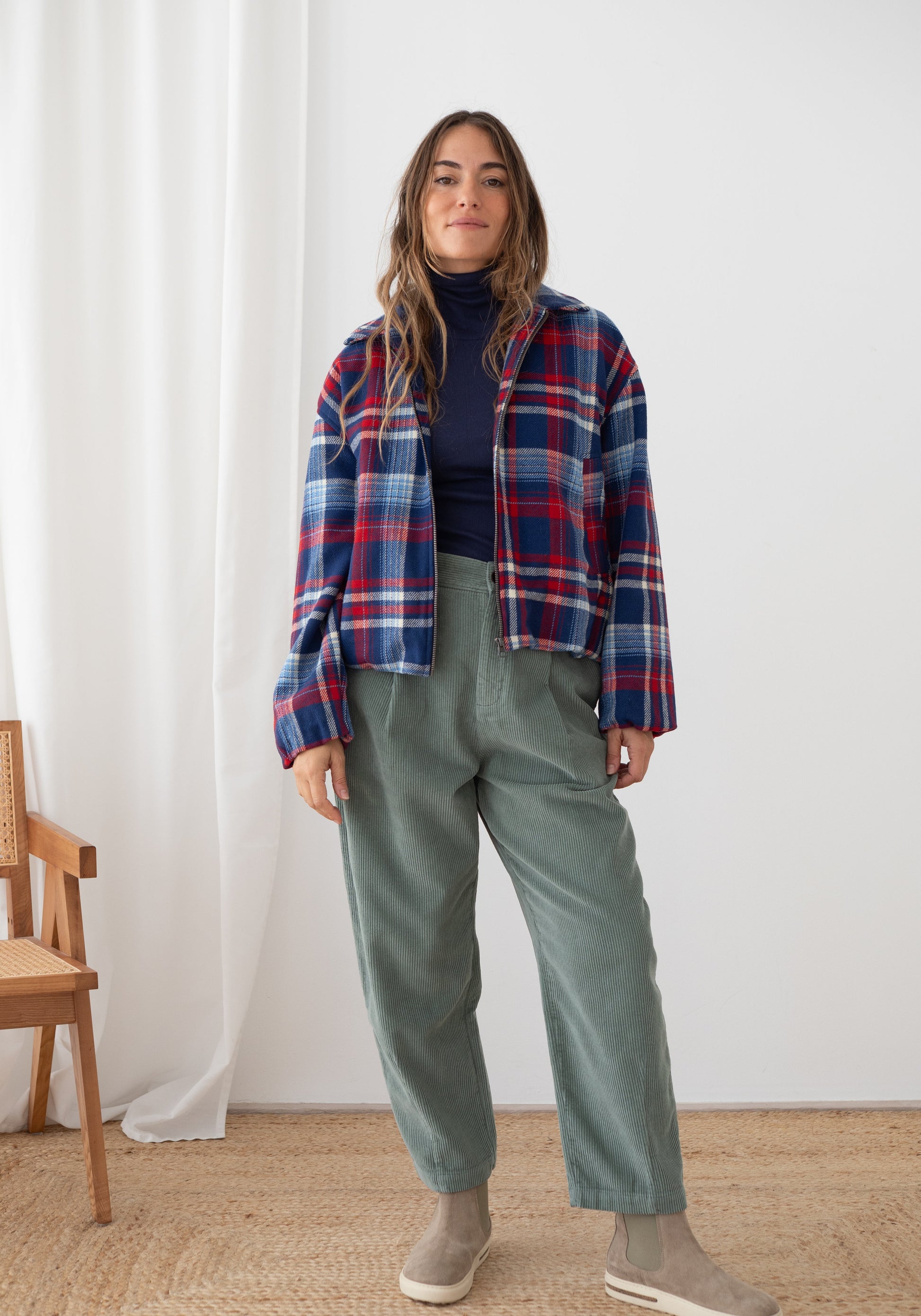 A woman stands indoors wearing a blue, red, and white plaid button-up shirt over a dark navy turtleneck, paired with sage green wide-leg pants and gray slip-on shoes. She is posed in front of a white curtain and wall, with a wooden chair and woven rug partially visible.