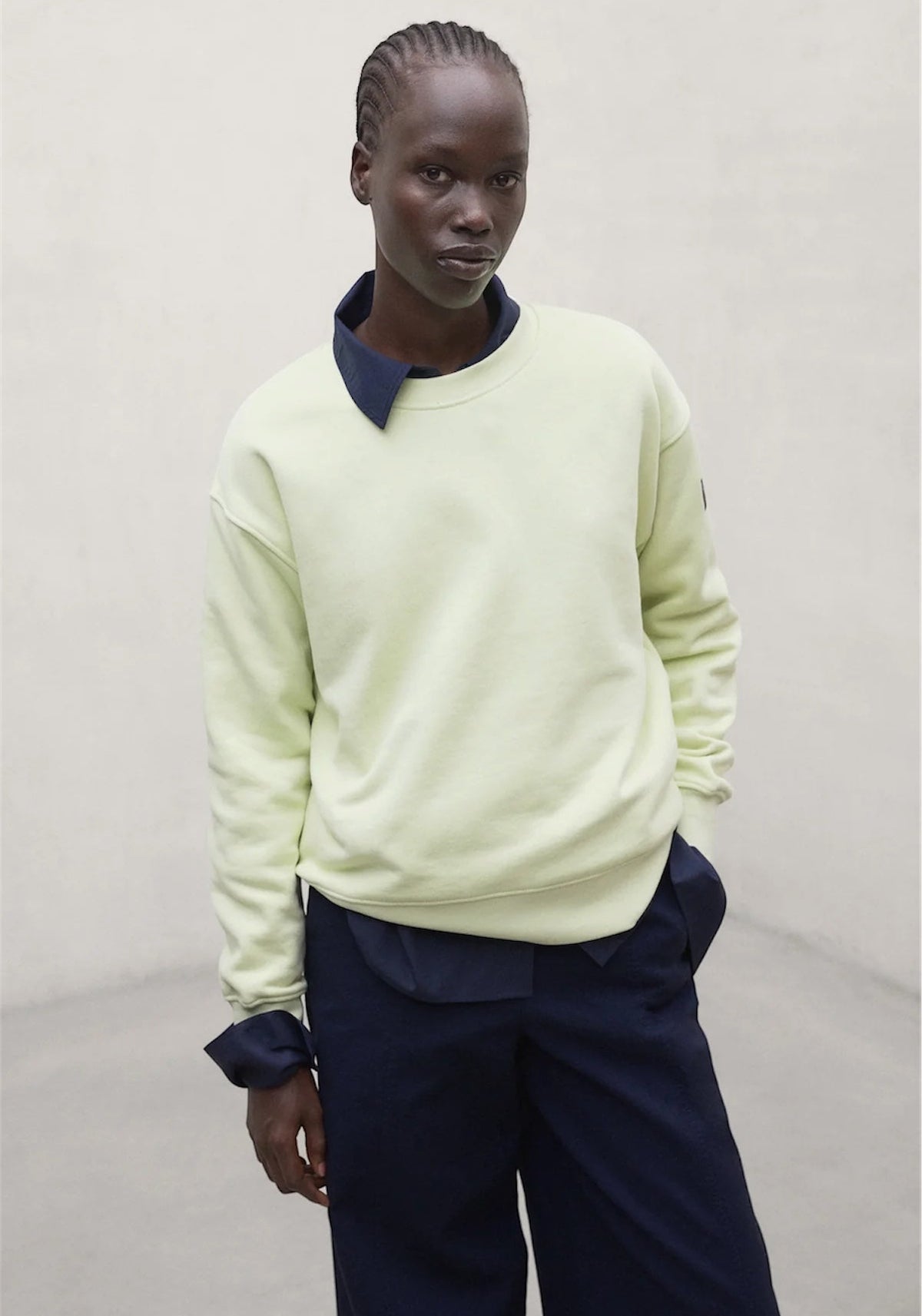 A model wearing a pale lime green crewneck sweatshirt from Ecoalf with a contrasting dark navy collar, paired with dark navy trousers, against a plain light gray background.