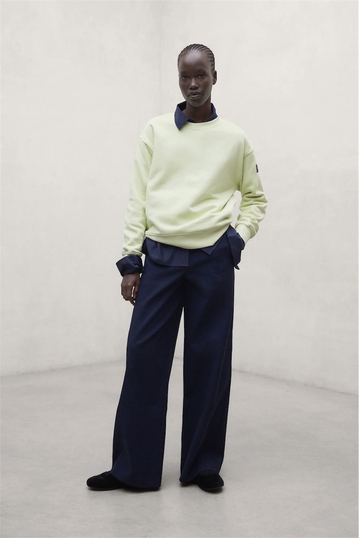 A model stands in a minimalist studio setting wearing a pale lime green long-sleeve sweatshirt by. ECOALF with dark navy blue collar and cuff details, paired with dark navy wide-leg trousers and black shoes.