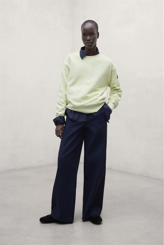 A model stands in a minimalist studio setting wearing a pale lime green long-sleeve sweatshirt by. ECOALF with dark navy blue collar and cuff details, paired with dark navy wide-leg trousers and black shoes.