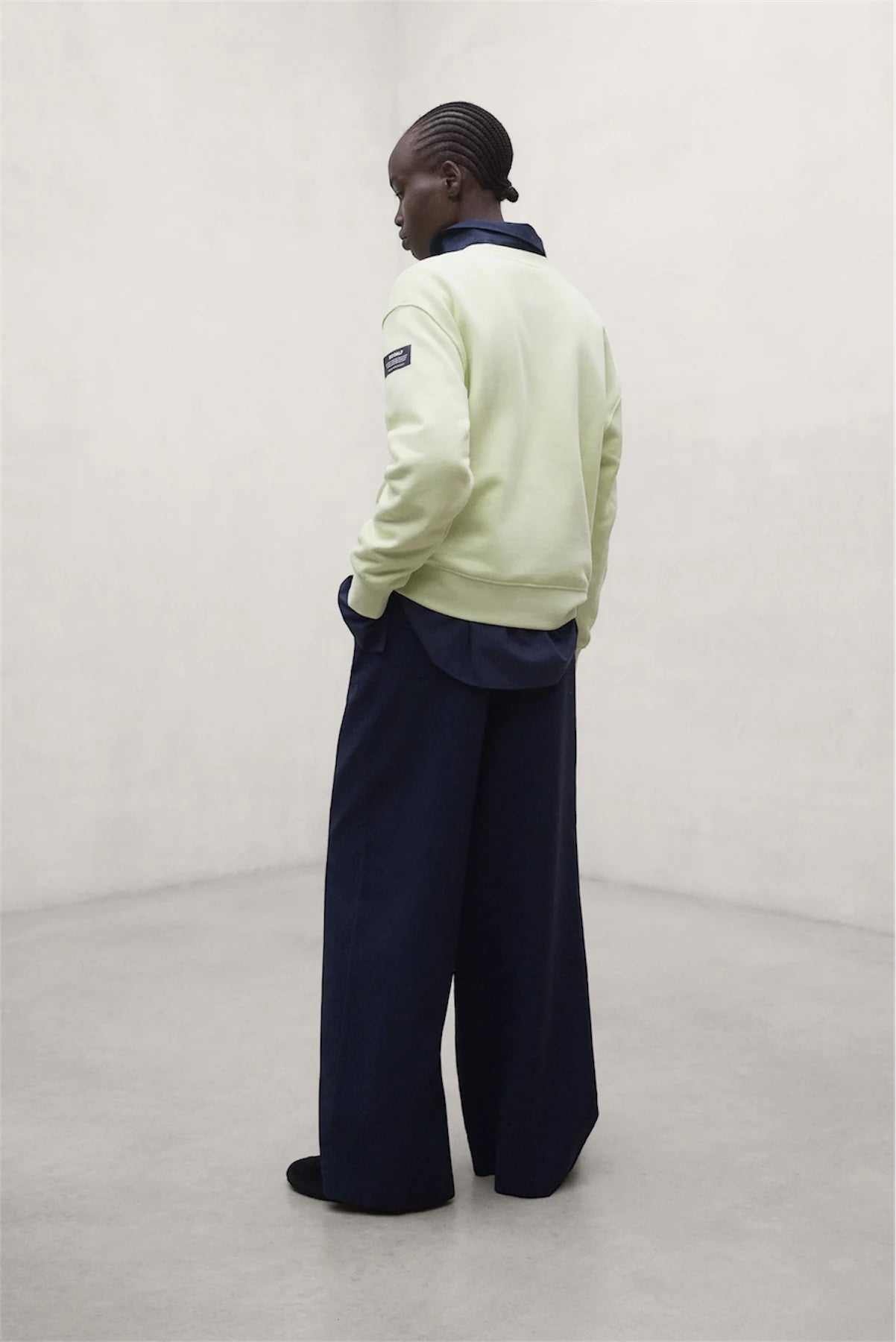 A model shown from the back wearing a pale yellow-green crewneck sweatshirt with a dark navy collar and sleeve patch, paired with wide-leg navy trousers, in a minimalist studio setting.