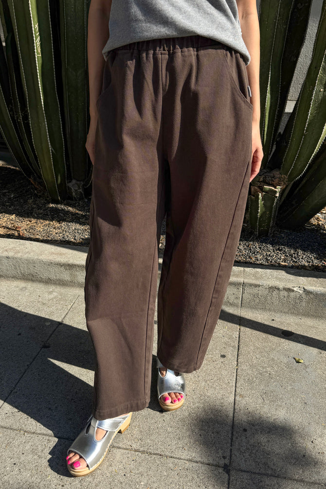 A person wearing loose brown wide-leg Arc pants by Le Bon Shoppe with a gray top and metallic silver sandals, standing outdoors on a concrete sidewalk in front of a cactus plant.