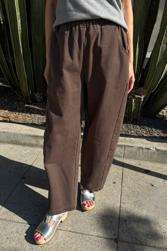 A person wearing loose brown wide-leg Arc pants by Le Bon Shoppe with a gray top and metallic silver sandals, standing outdoors on a concrete sidewalk in front of a cactus plant.