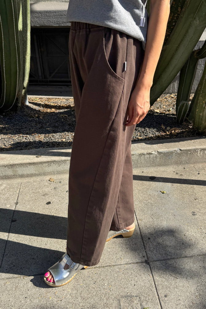 A person wearing loose brown wide-leg Arc pants by Le Bon Shoppe, a gray top, and metallic silver slide sandals, standing outdoors on a concrete sidewalk near cacti.