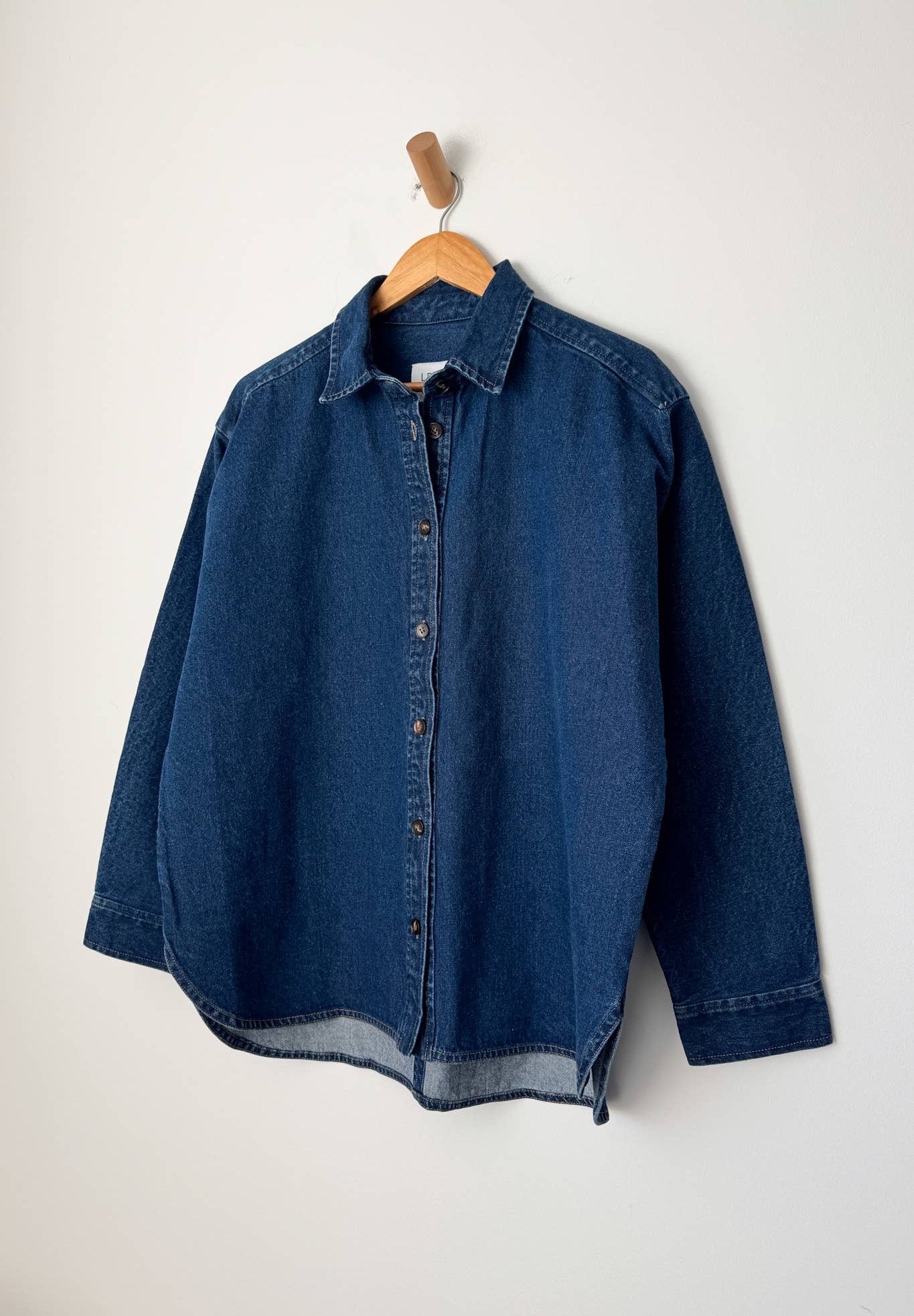 A dark blue cotton denim shirt from Le Bon Shoppe hangs on a wooden hanger against a plain white wall. The shirt features a classic button-down front, a small collar, and long sleeves with buttoned cuffs, showcasing a slightly relaxed fit. A wooden wall hook is visible above the hanger, adding a minimalist touch to the display.

