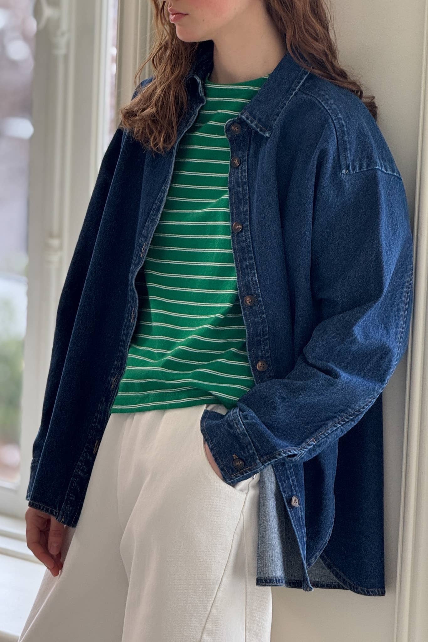A woman with long, wavy hair stands near a window, wearing a dark blue cotton denim shirt from Le Bon Shoppe. The shirt features a button-down front, a small collar, and long sleeves, layered over a green and white striped shirt, and paired with relaxed white trousers. The casual, slightly oversized fit of the denim shirt creates a laid-back yet stylish look against the soft natural light coming through the window.

