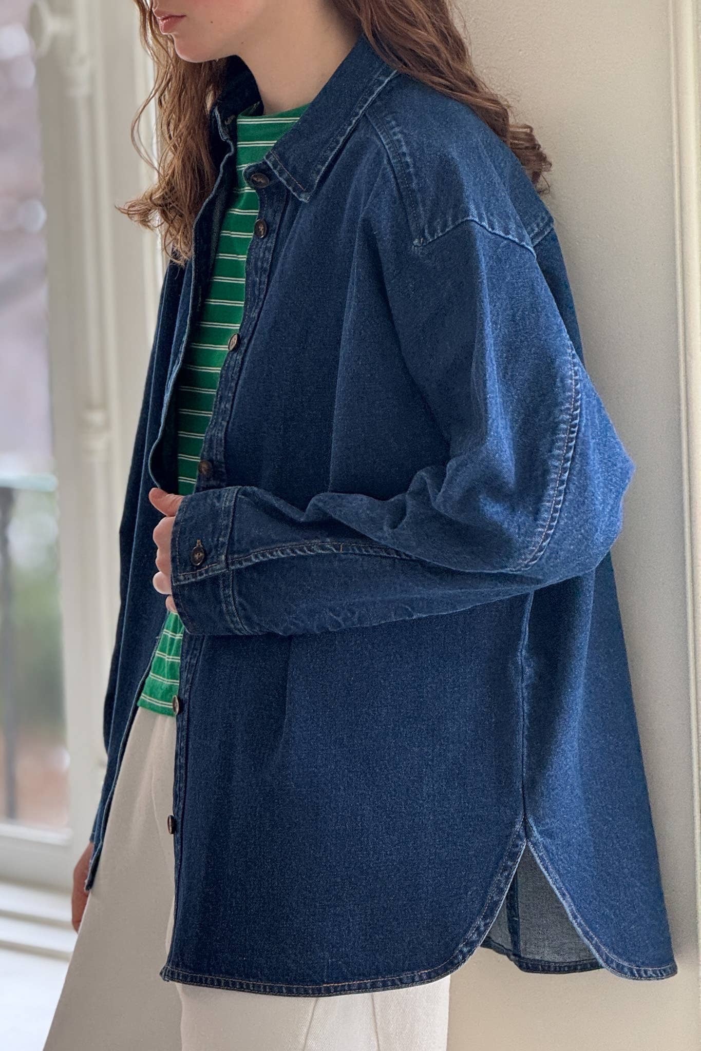 A woman with long, wavy hair stands near a window, wearing a dark blue cotton denim shirt from Le Bon Shoppe. The shirt features a button-down front, a small collar, and long sleeves with buttoned cuffs, layered over a green and white striped shirt, and paired with relaxed white trousers. The slightly oversized denim shirt creates a casual, effortless look, with natural light from the window softly illuminating the scene.

