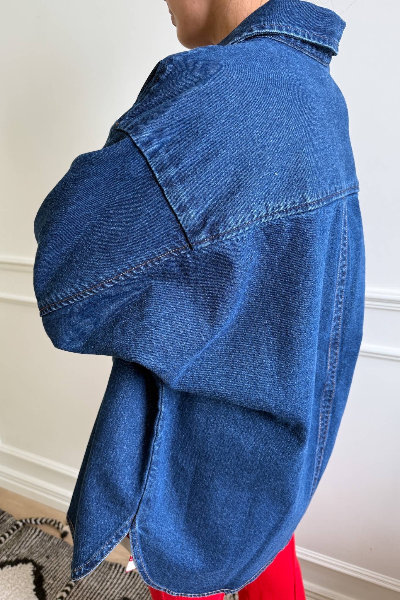 A person stands indoors, viewed from the back, wearing a dark blue cotton denim shirt from Le Bon Shoppe. The shirt features a small collar, long sleeves, and a slightly oversized fit, paired with bright red trousers. The scene includes a white wall with decorative molding and a patterned rug on the floor, adding a cozy backdrop to the casual outfit.

