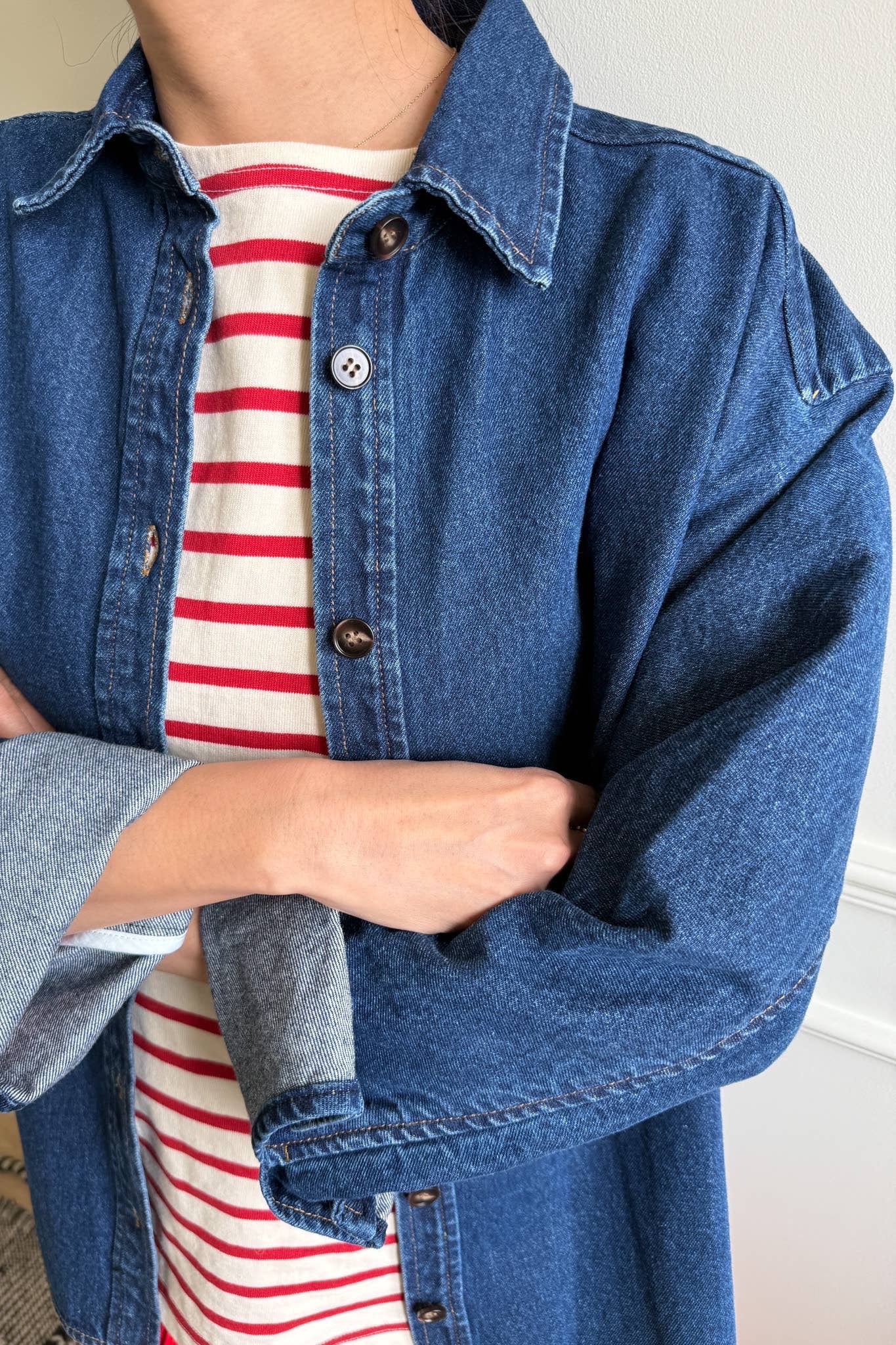 A close-up of a person wearing a dark blue cotton denim shirt from Le Bon Shoppe, featuring a button-down front, a small collar, and long sleeves with rolled-up cuffs. The shirt is layered over a red and white striped top, with the person's arms crossed, highlighting the relaxed, slightly oversized fit. The background includes a white wall with decorative molding and a patterned rug on the floor, adding a cozy setting to the outfit.

