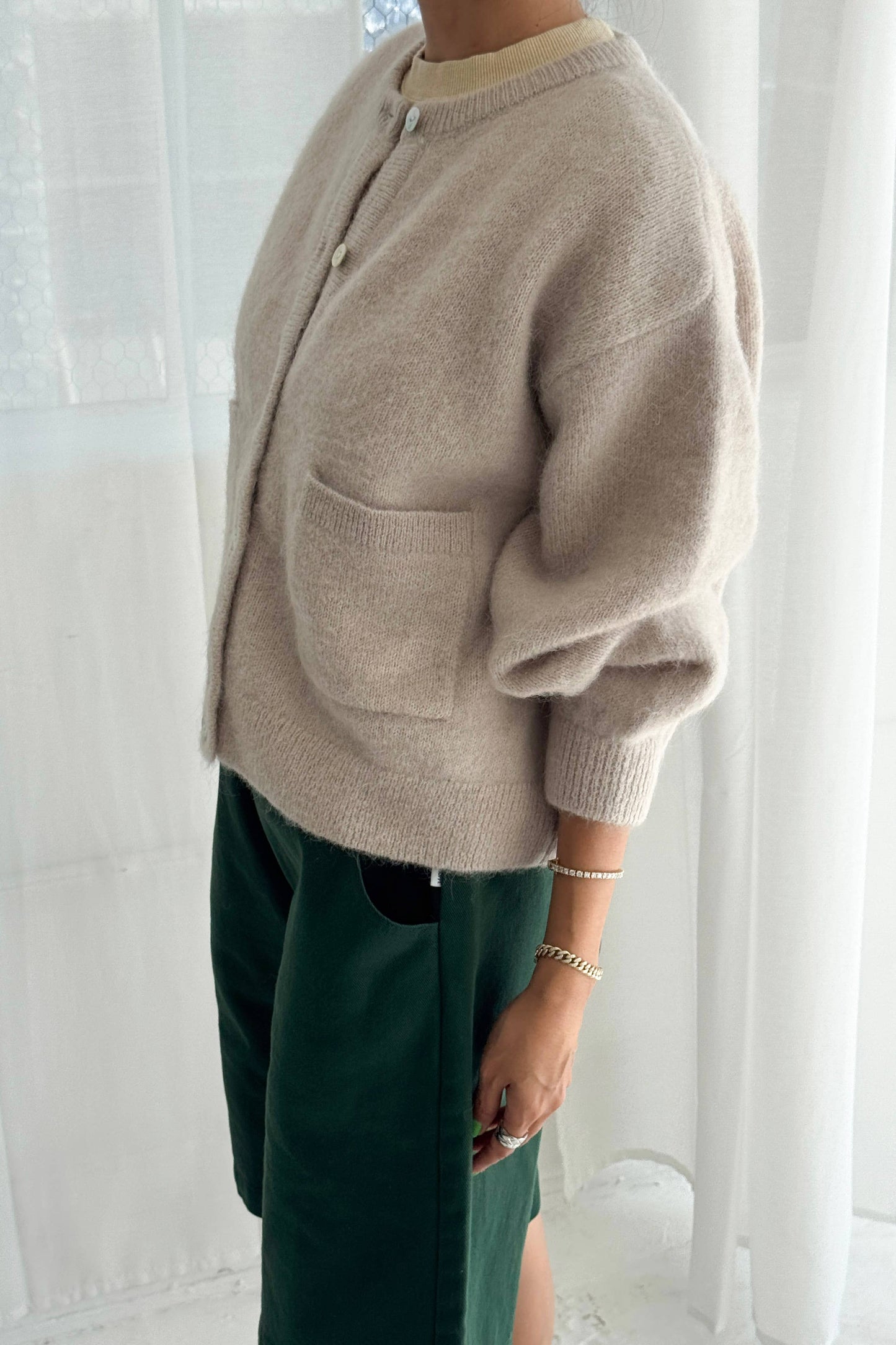 Person wearing a beige Amelie cardigan from Le Bon Shoppe and green pants against a white curtain background