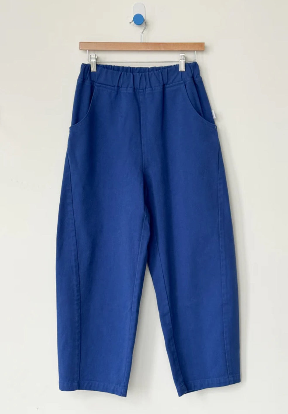 Azure blue Arc pants from Le Bon Shoppe hanging on a wooden hanger against a white background
