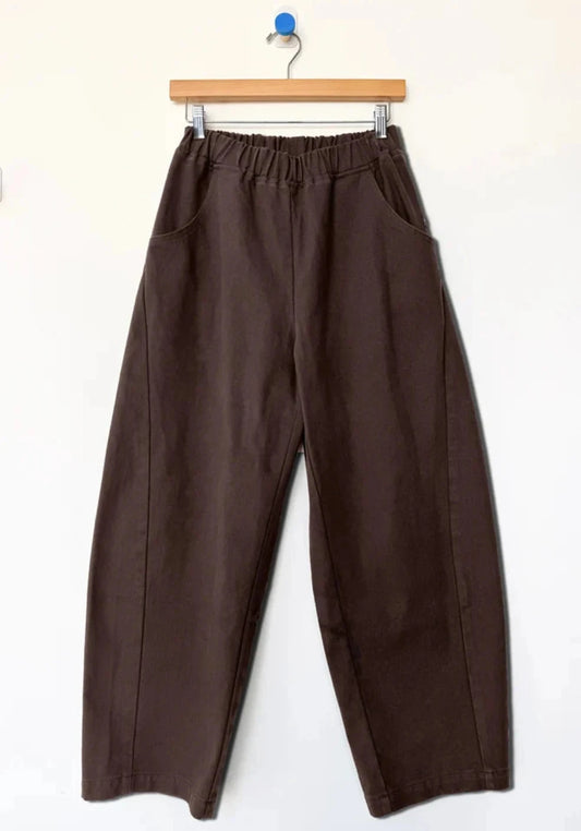 Pair of wide-leg cropped barrel leg Arc pants by Le Bon Shoppe in a dark chocolate brown color, hanging from a wooden hanger against a plain white wall.
