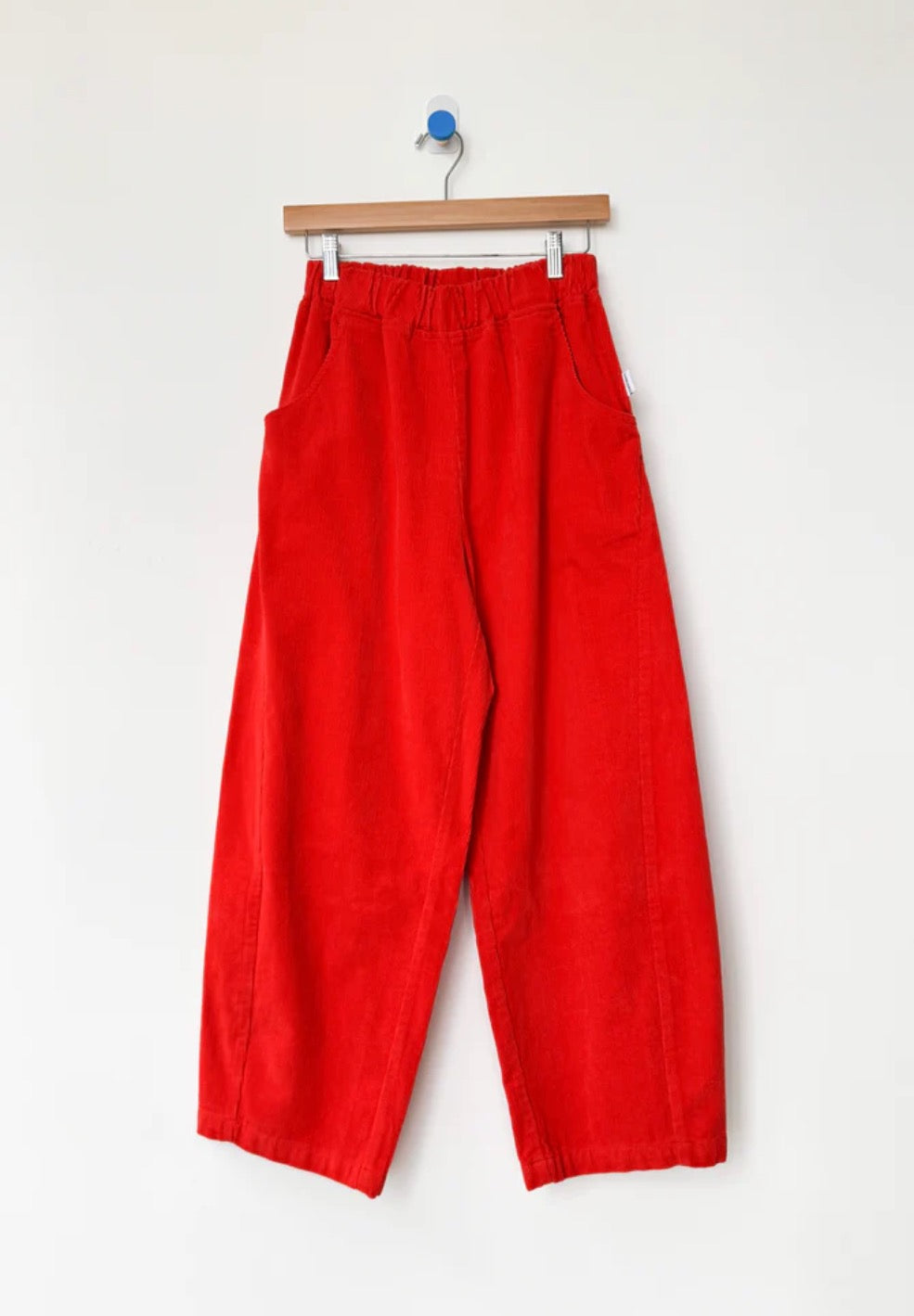 Bright red corduroy Arc pants from Le Bon Shoppe hanging on a wooden hanger against a white background