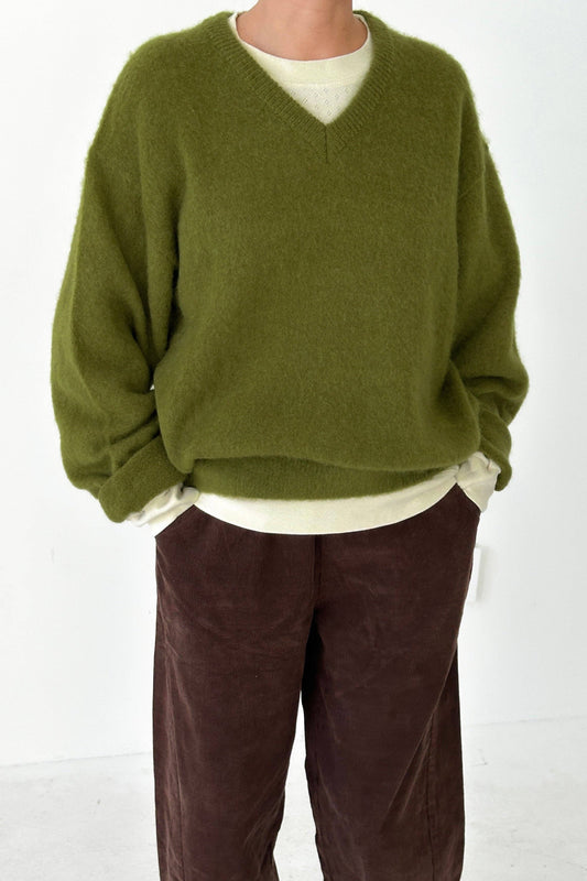 Person wearing a leaf green mohair James sweater from Le Bon Shoppe over a white shirt with brown pants on a white background