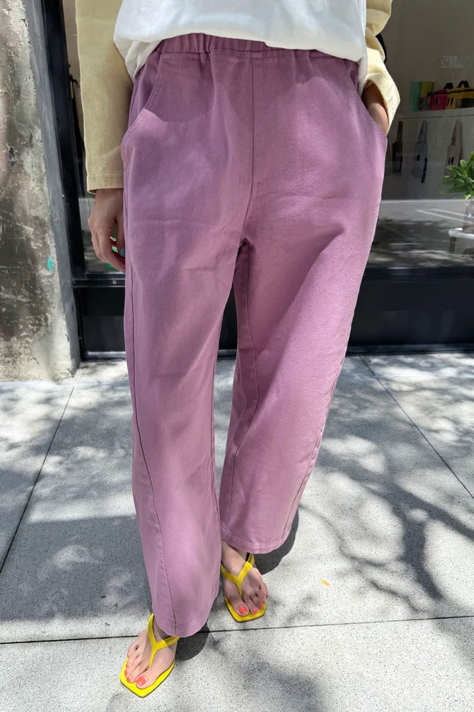 A person wearing loose, wide-leg mauve-purple Arc pants by Le Bon Shoppe with a white top and bright yellow sandals, standing outdoors on a paved walkway.