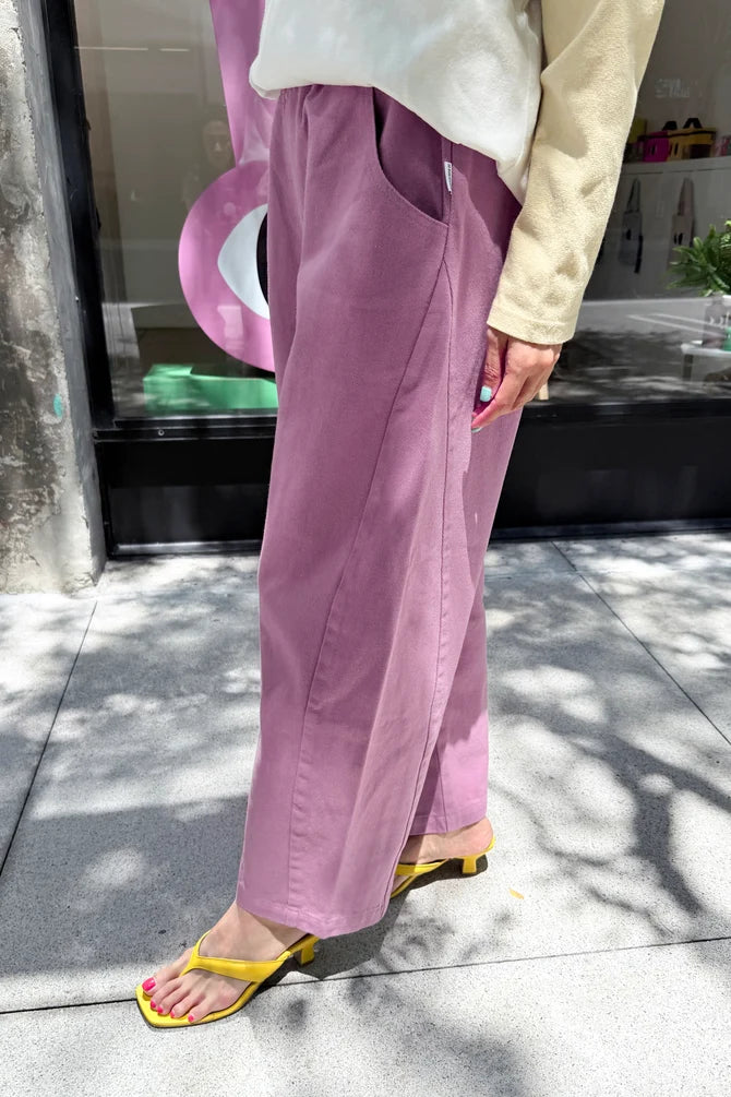 A person outdoors wearing wide-leg mauve-purple Arc pants by Le Bon Shoppe, a cream long-sleeve top, and yellow heeled sandals, standing on a paved walkway in front of a storefront.