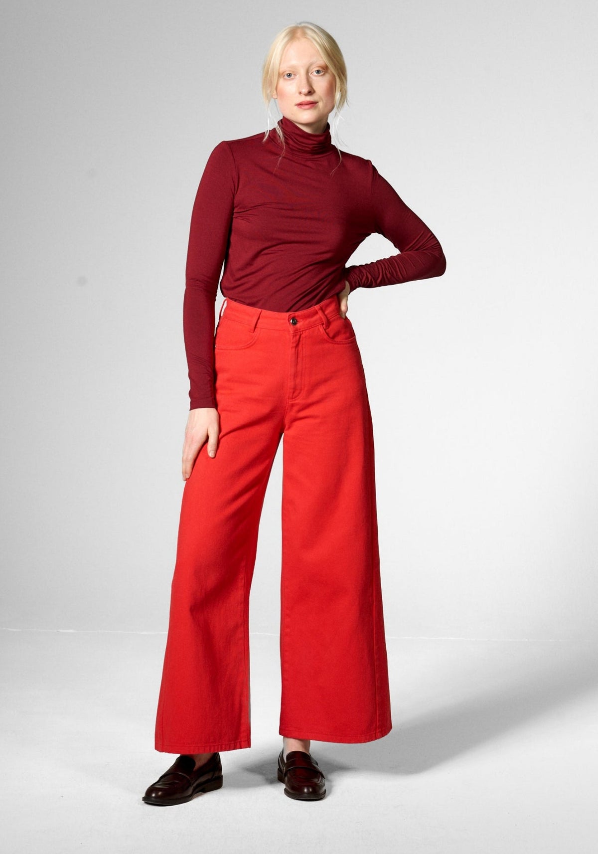A model stands against a light gray studio background wearing a dark red long-sleeve turtleneck top and bright red wide-leg jeans by Mademoiselle Yeye, paired with dark brown leather shoes.