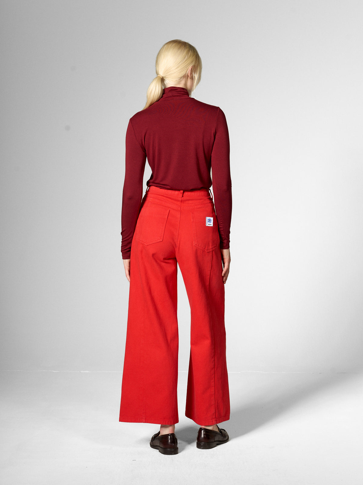 A woman viewed from behind wearing a dark red mock-neck long-sleeve top and bright red wide-leg trousers by Mademoiselle Yeye, standing against a light grey studio background.