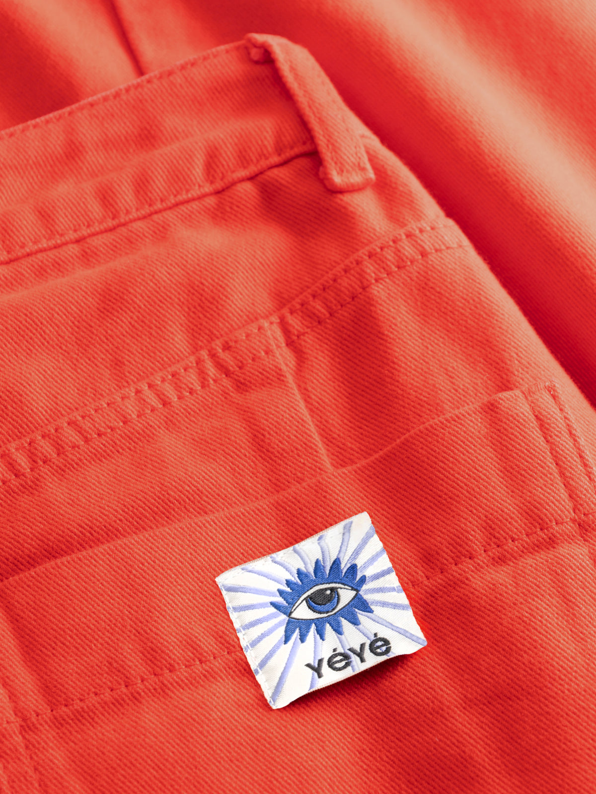 Close-up of bright orange denim fabric showing the back pocket and a small square Yèvé logo patch with a blue eye design.