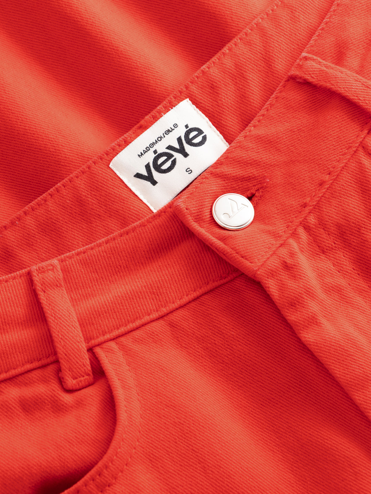 Close-up of bright orange-red denim fabric showing a white Yèvé label and a white button at the waistband.