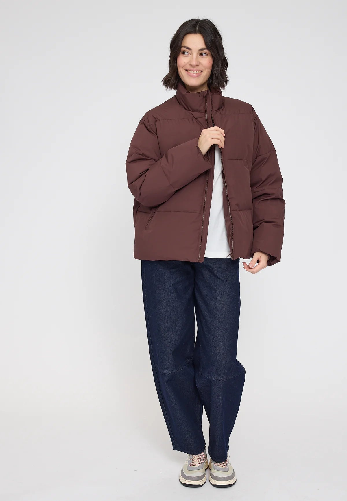 Woman wearing a chocolate brown puffer jacket and blue barrel leg jeans on a white background