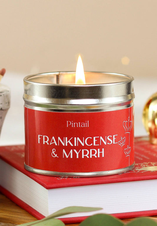 Candle in a red tin labeled 'Frankincense & Myrrh' by Pintail, lit on a surface with a blurred background.