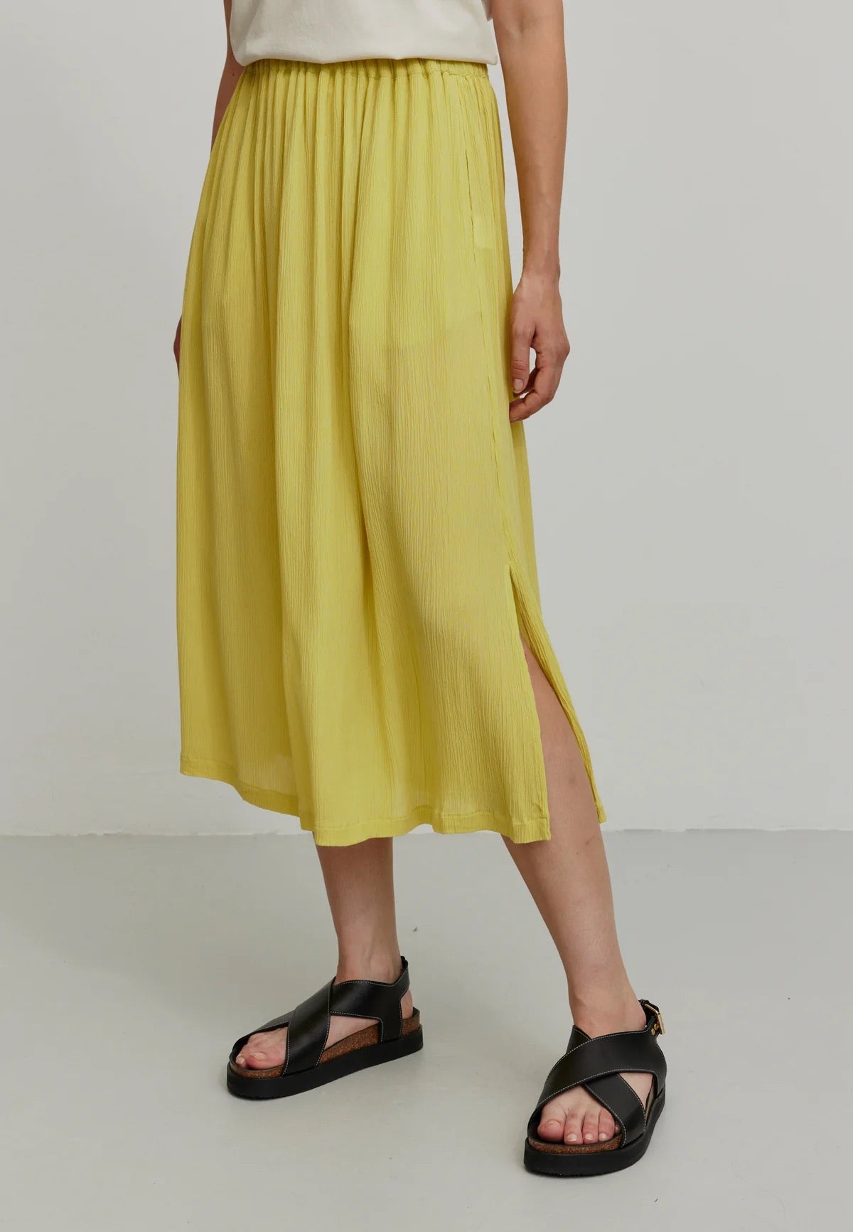 A person models a vibrant mustard yellow LENZING™ ECOVERO™ midi skirt from Recolution, featuring a high-waisted, elastic waistband with a gathered design and a subtle side slit. The skirt has a soft, crinkled texture and flows gracefully, paired with a white short-sleeve t-shirt and black strappy sandals. The person stands against a plain light gray background, with one hand in the skirt's side pocket and the other resting by their side.

