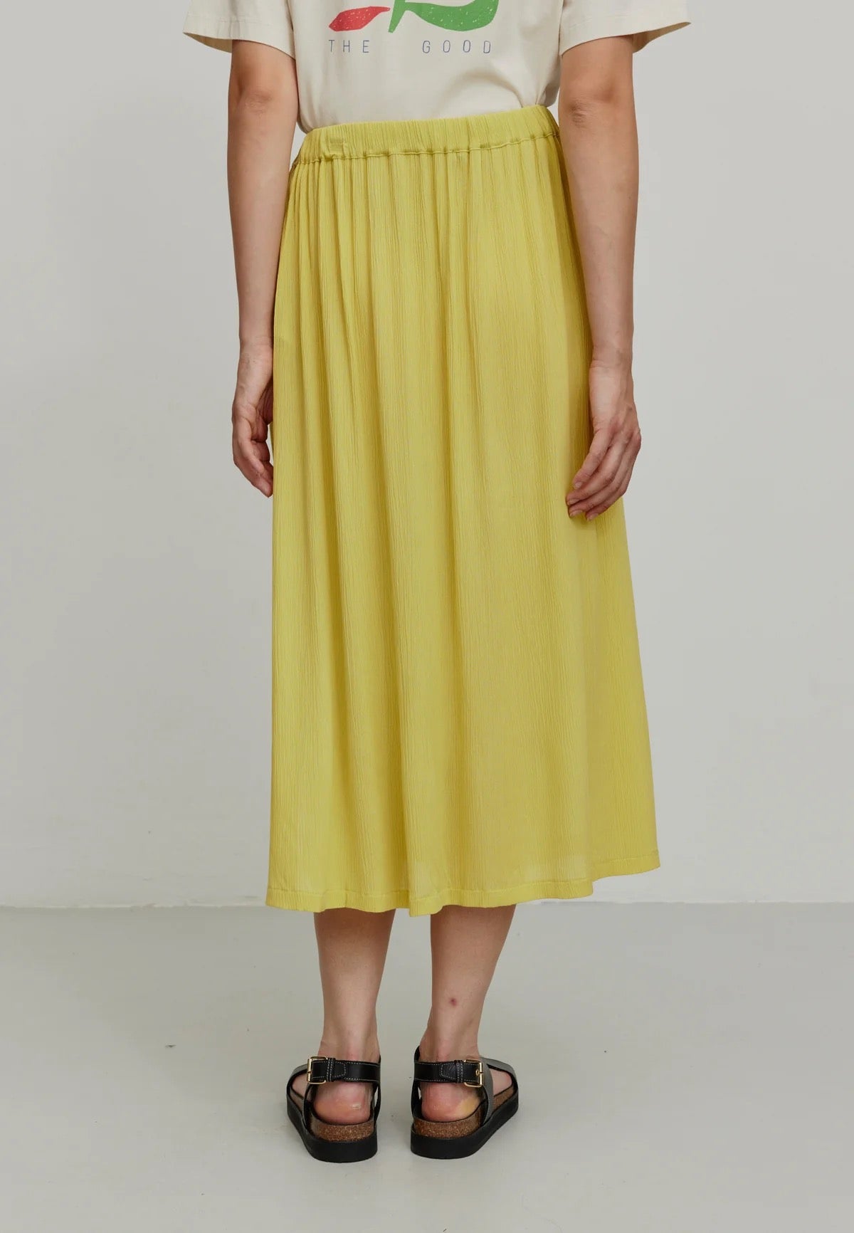 A person models a vibrant mustard yellow LENZING™ ECOVERO™ midi skirt from Recolution, featuring a high-waisted, elastic waistband with a gathered design and a subtle side slit. The skirt has a soft, crinkled texture and flows gracefully, paired with a white short-sleeve t-shirt and black strappy sandals. The person stands against a plain light gray background, with one hand in the skirt's side pocket and the other resting by their side.

