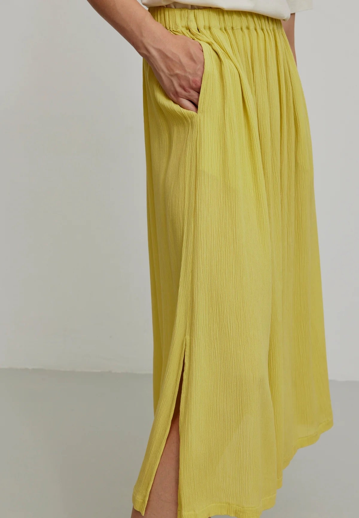 A person models a vibrant mustard yellow LENZING™ ECOVERO™ midi skirt from Recolution, featuring a high-waisted, elastic waistband with a gathered design and a subtle side slit. The skirt has a soft, crinkled texture and flows gracefully, paired with a white short-sleeve t-shirt and black strappy sandals. The person stands against a plain light gray background, with one hand in the skirt's side pocket and the other resting by their side.

