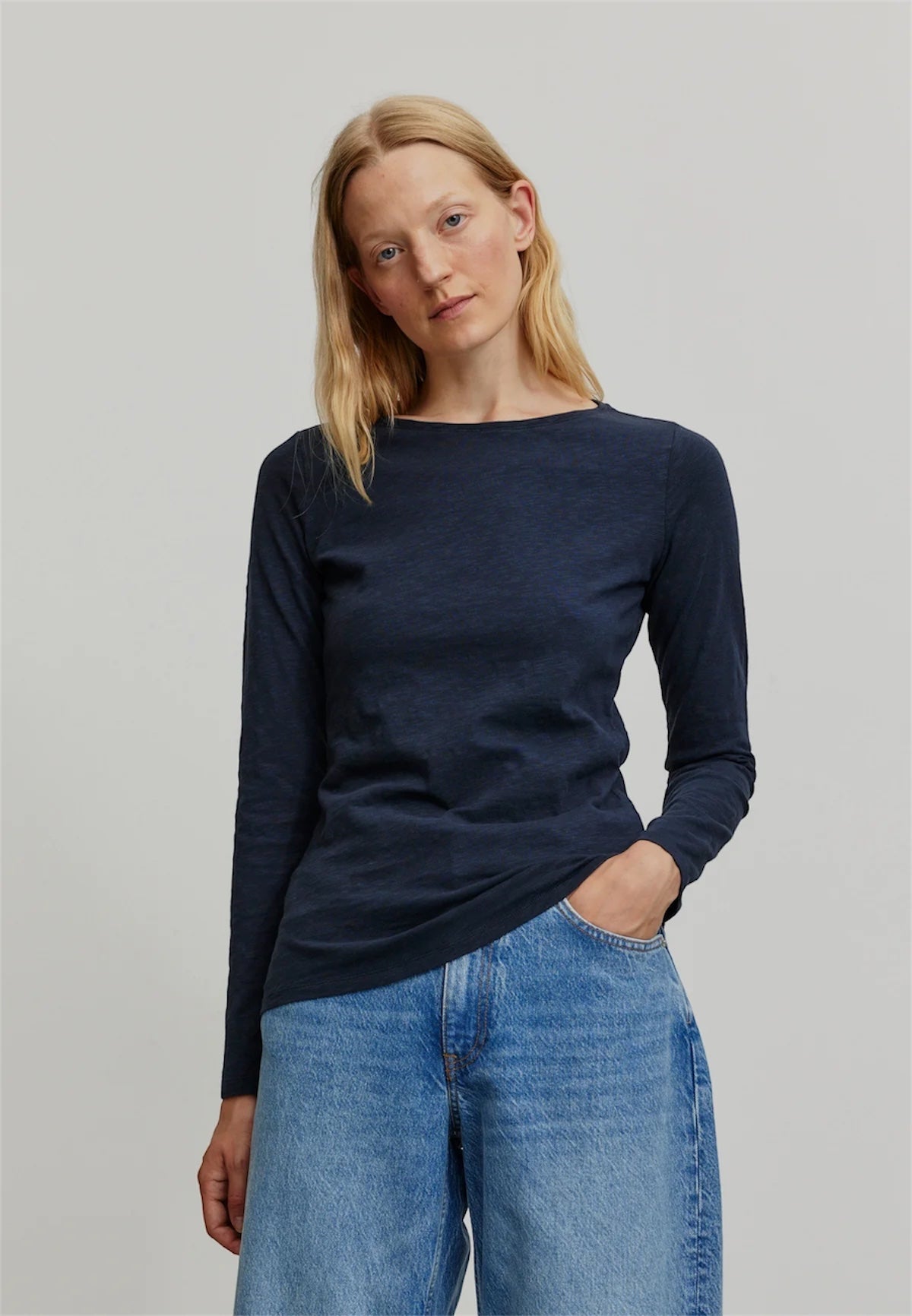 A woman modeling a dark navy blue organic cotton long-sleeve boat-neck top from Recolution with light blue jeans, standing against a neutral light gray studio background.
