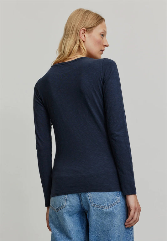 A woman shown from the back wearing a dark navy organic cotton long-sleeve slub jersey top from Recolution and light blue jeans against a plain light gray studio background.