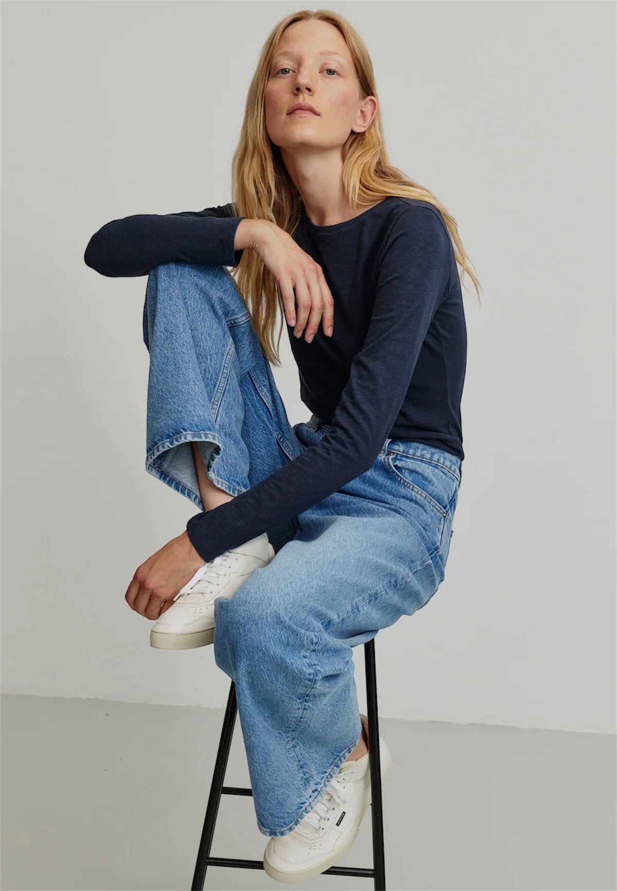 Woman sitting on a stool wearing a navy organic cotton long-sleeve top by Recolution, blue jeans, and white sneakers.