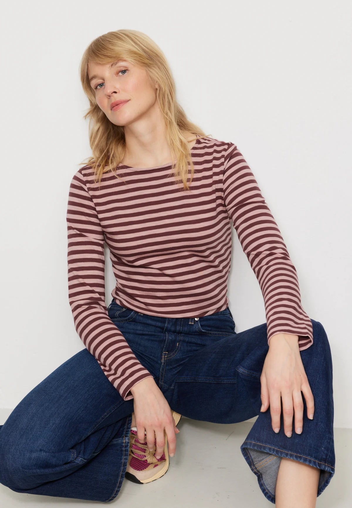 Woman wearing a striped long-sleeve top and blue jeans sitting on the floor against a white background